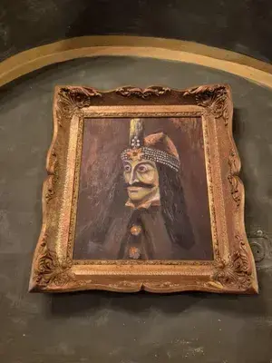 Ritratto in cornice dorata di Vlad l’Impalatore con corona ingioiellata e tunica scura, appeso a una parete scura.